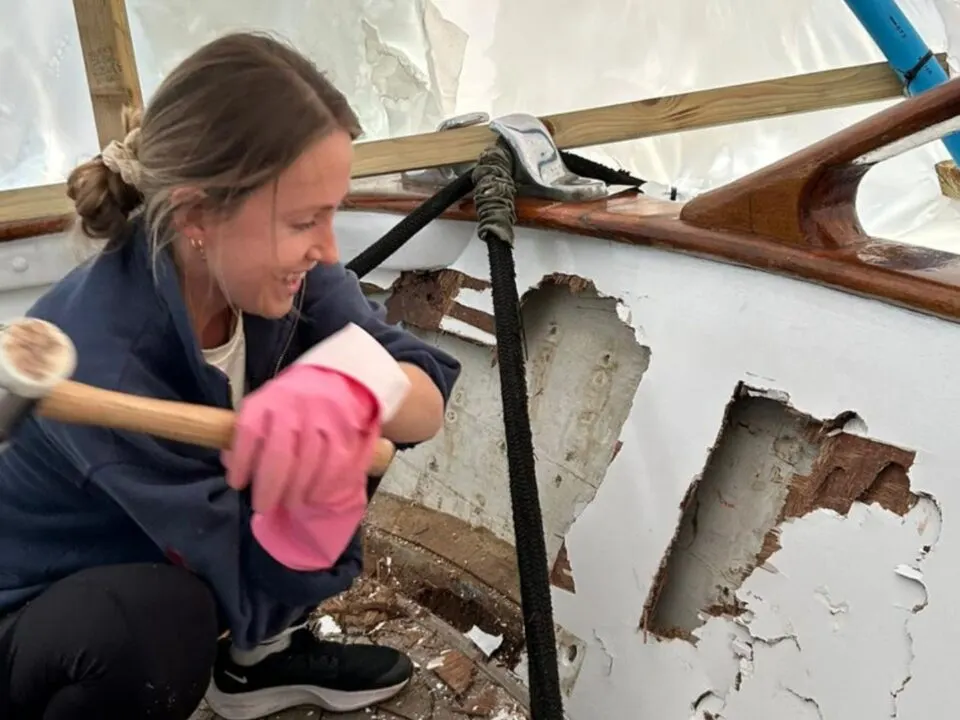 Woman scraping paint from wooden hull interior of a sailing vessel during maintenance work.