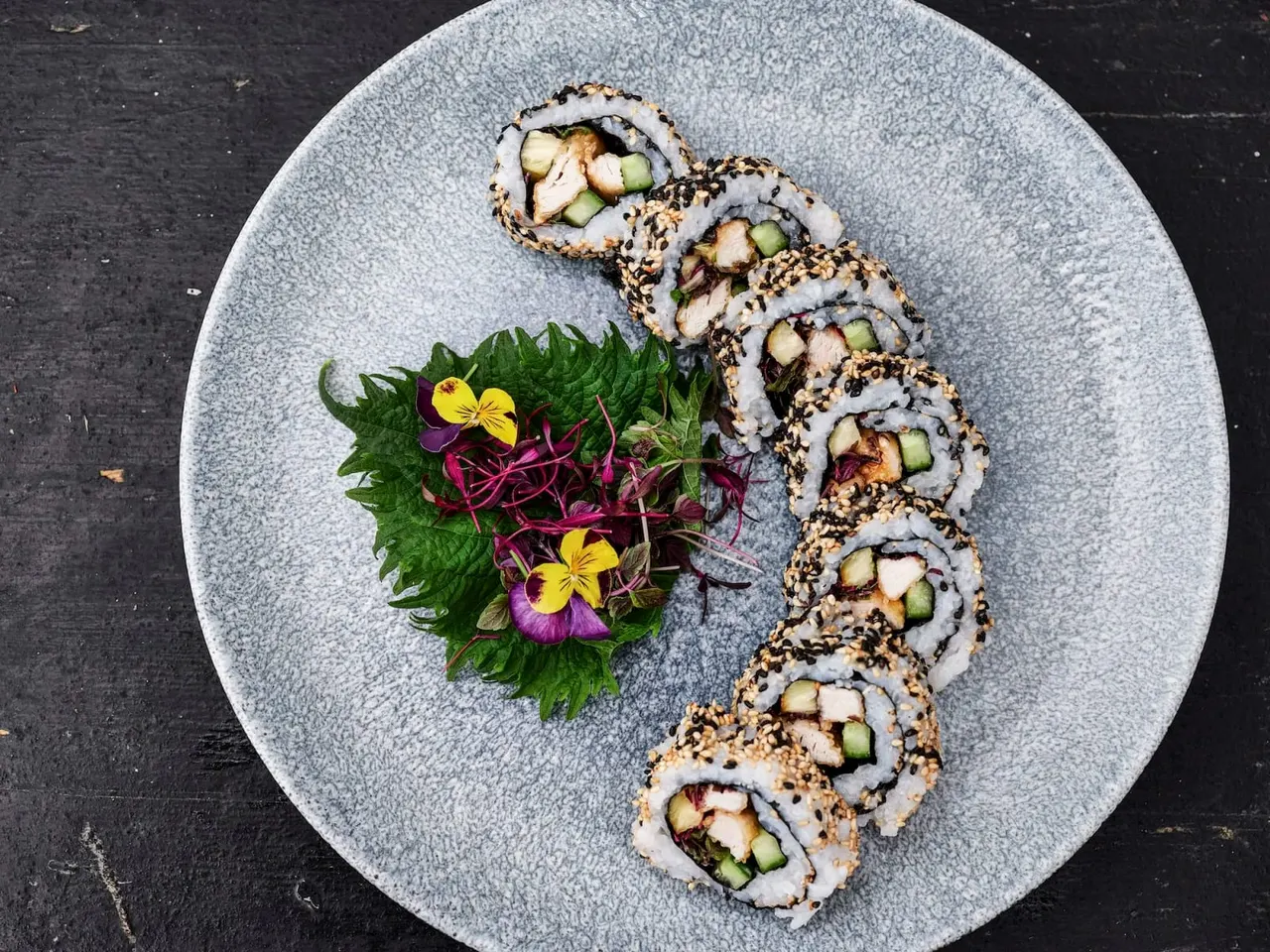 Sushi rolls with sesame seed coating arranged on a grey plate with edible flowers and microgreens.