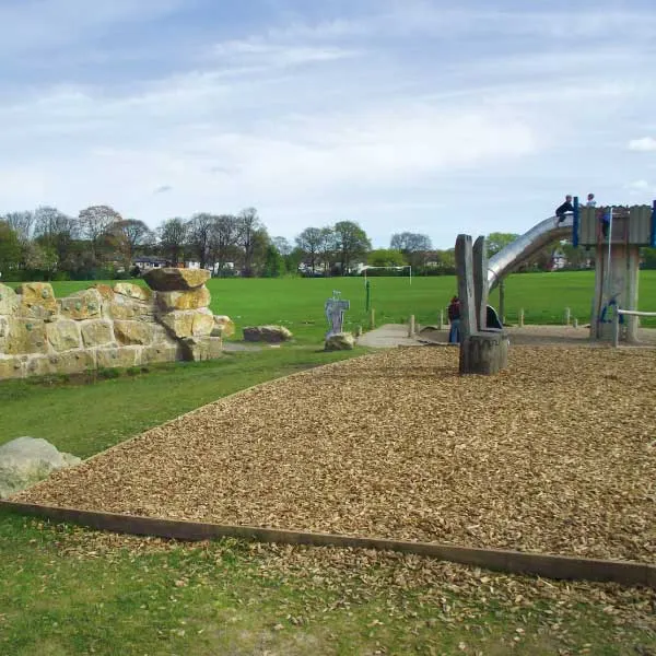 Outdoor play area with climbing structures, stone features and gravel ground overlooking green fields.