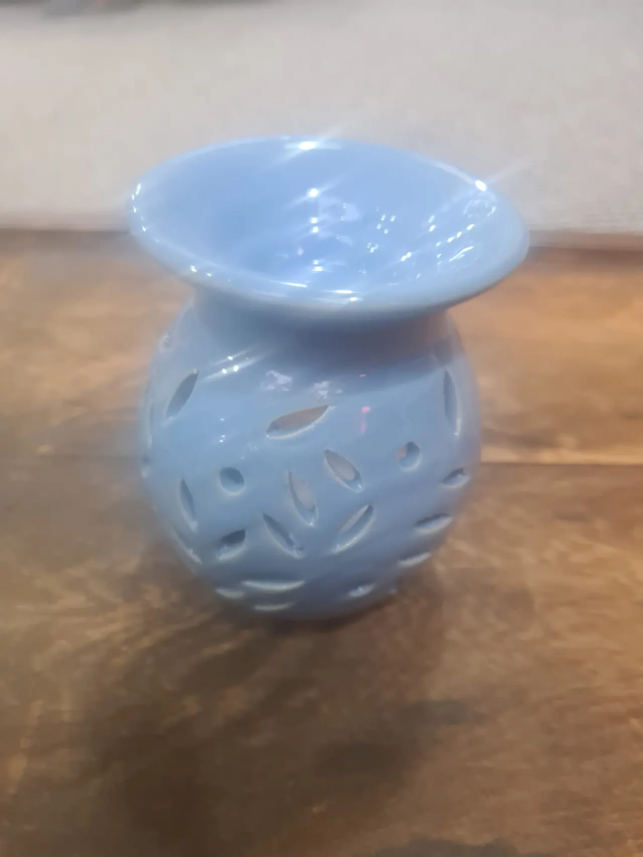 A small pale blue ceramic oil burner with a wide bowl top and perforated body on a wooden surface.