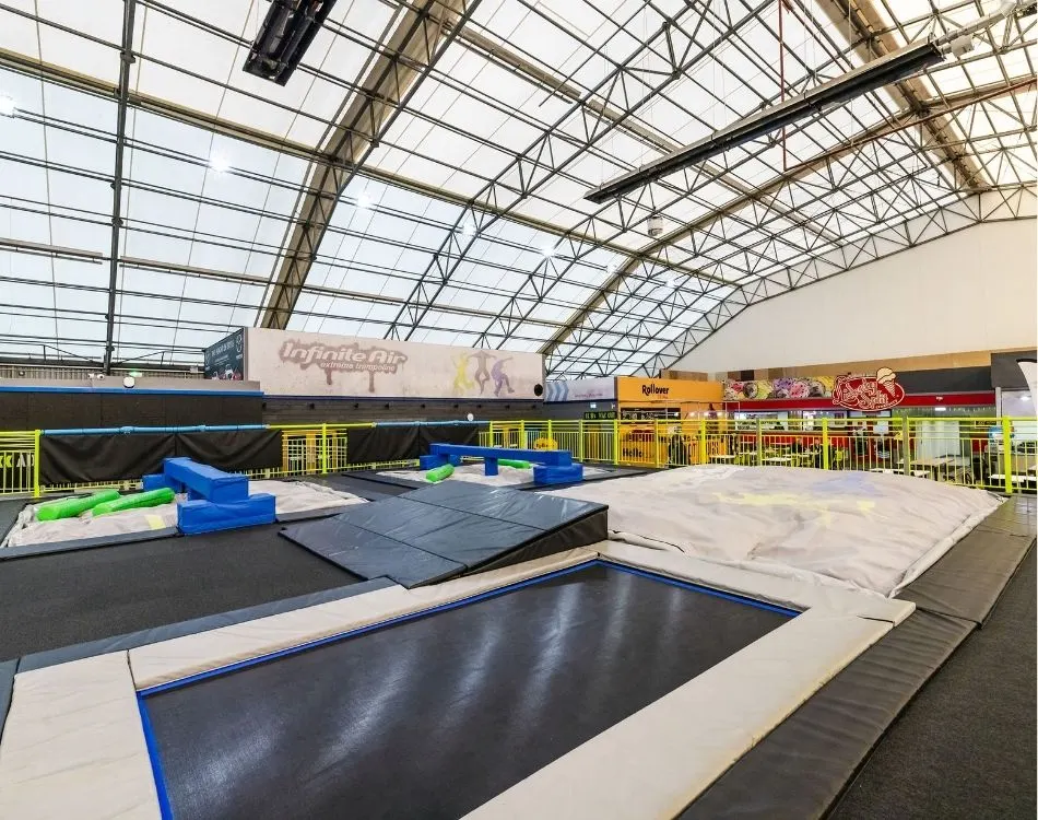 Indoor trampoline park with multiple black trampolines, foam pits and blue foam blocks under a glass ceiling.