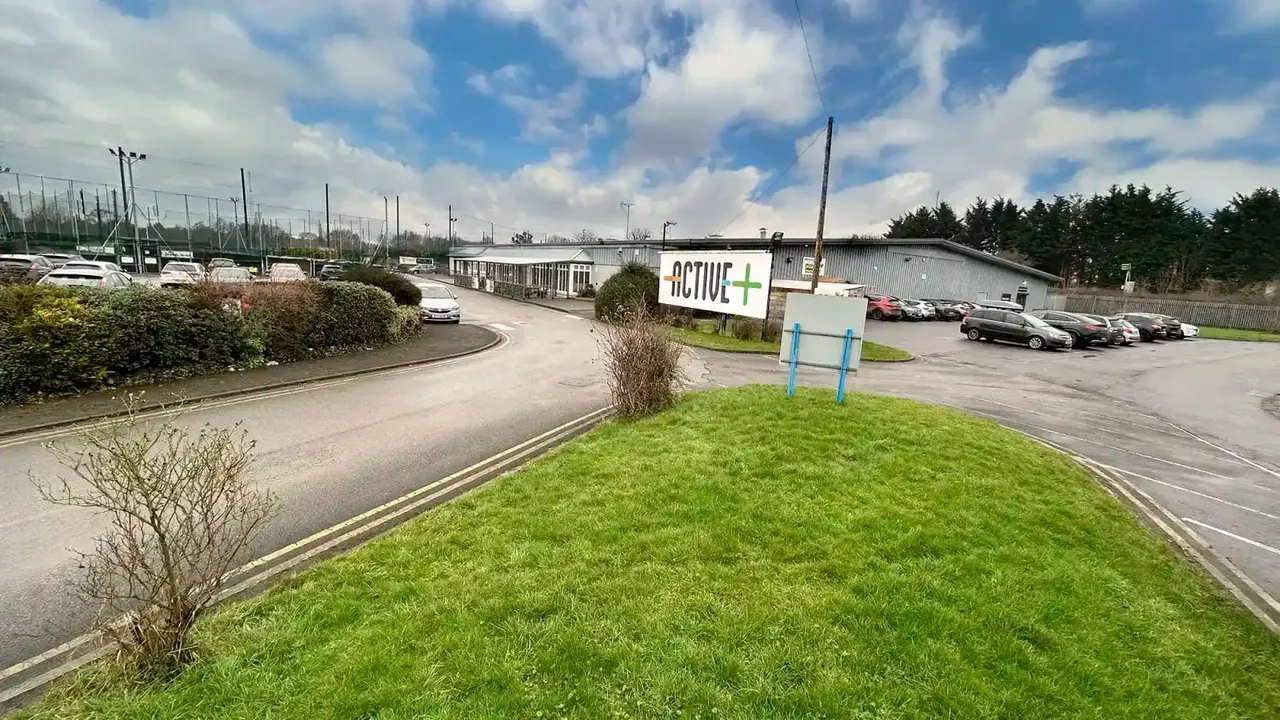 Sports facility grounds showing car park, buildings with Active+ signage, and tennis courts behind fencing.