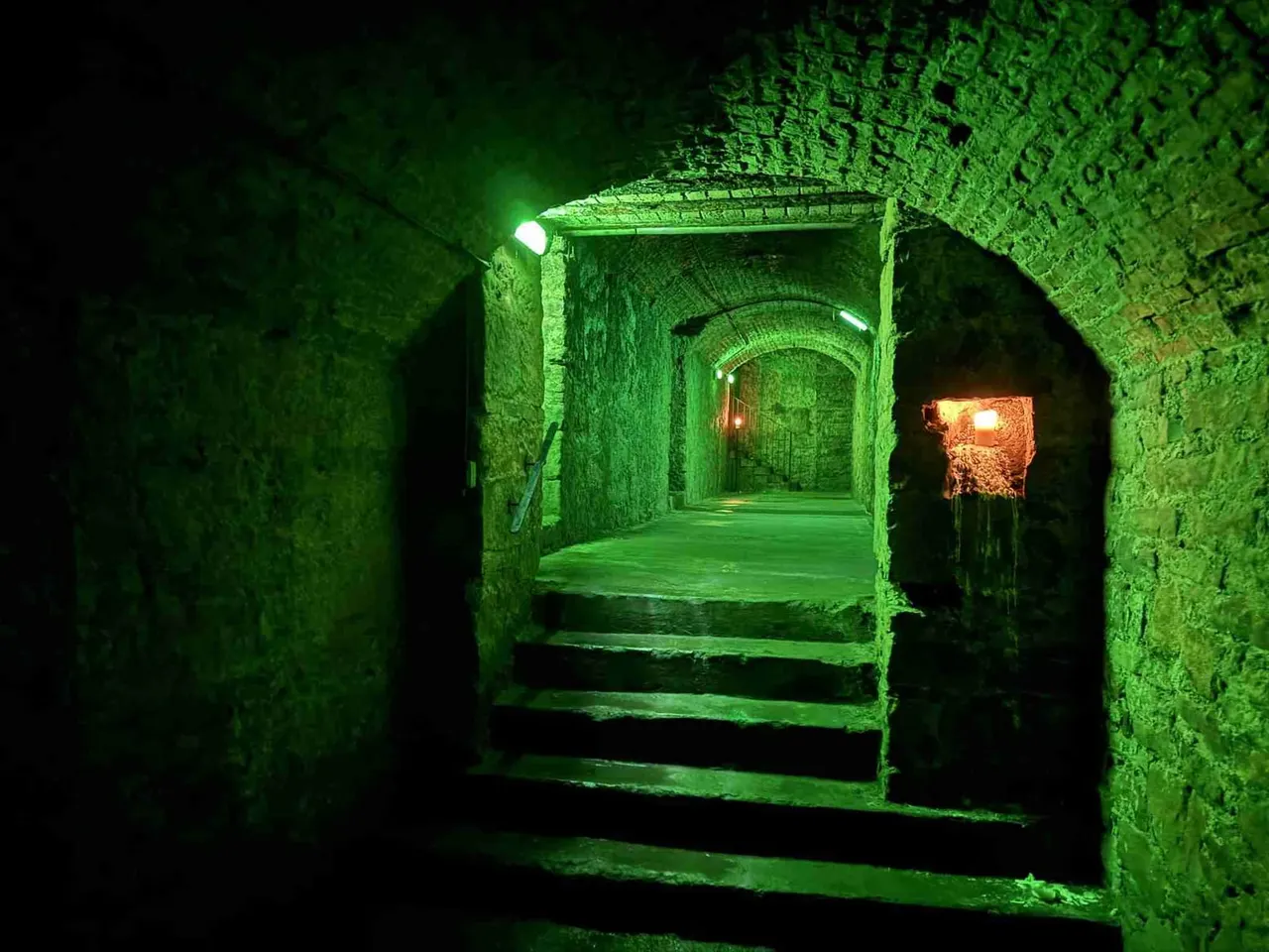 Historic underground brick tunnel lit in green, with stone steps leading deeper into darkness and a small fire visible in the distance.