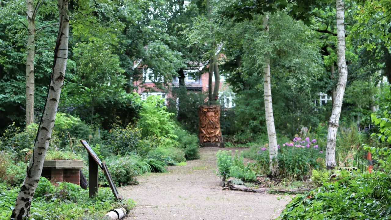 Wooded garden path with birch trees, flowering plants and a carved wooden sculpture in the distance.