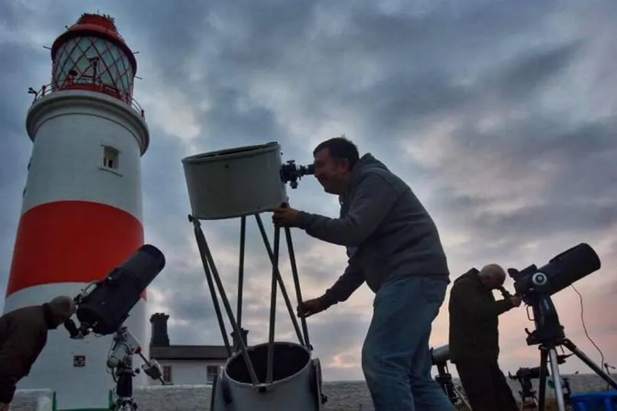 People using telescopes to observe the sky near a red and white lighthouse.
