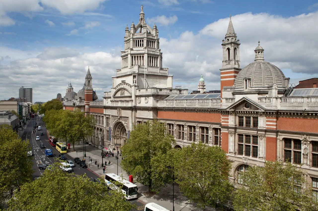 Historic London street with Victorian and Edwardian buildings, including museums and churches with towers, buses and pedestrians below.
