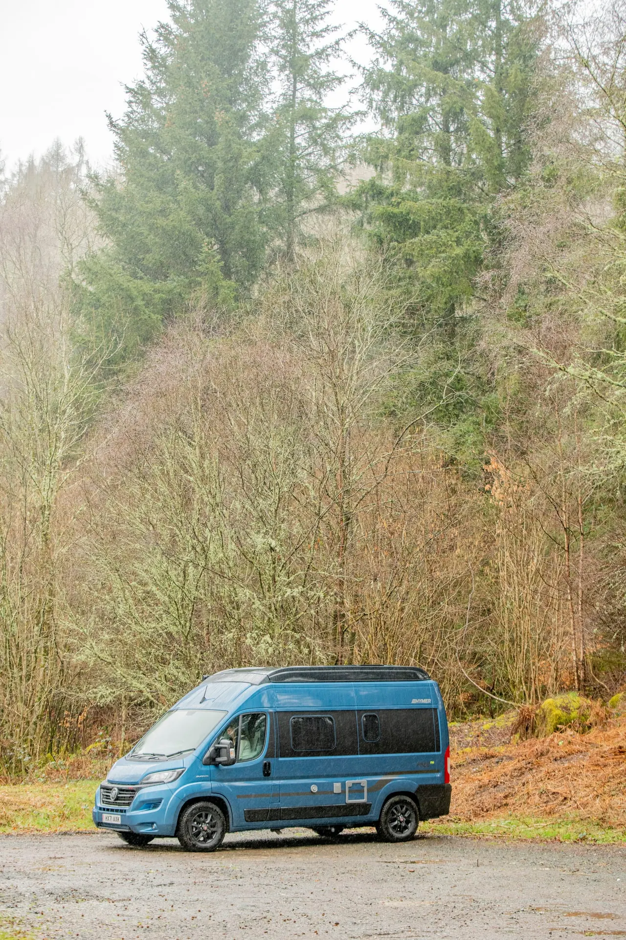 Blue campervan parked on gravel beside a woodland of bare and evergreen trees.