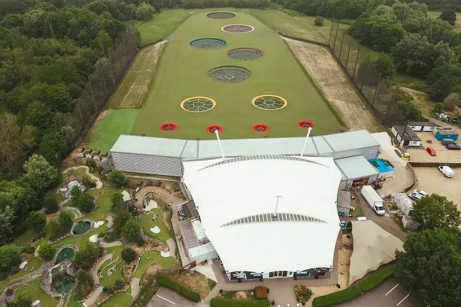 Aerial view of a golf entertainment venue with a long grass range lined with coloured target circles and red umbrellas, surrounded by woodland and facilities buildings.