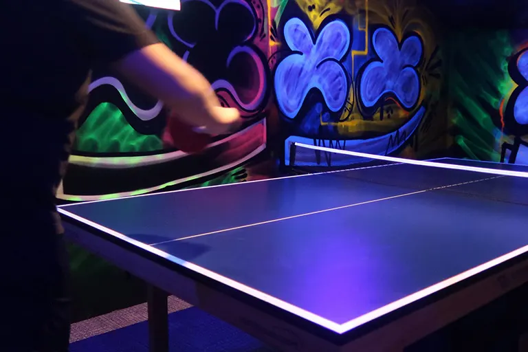 Person playing table tennis at an illuminated neon table in a dark arcade or entertainment venue.