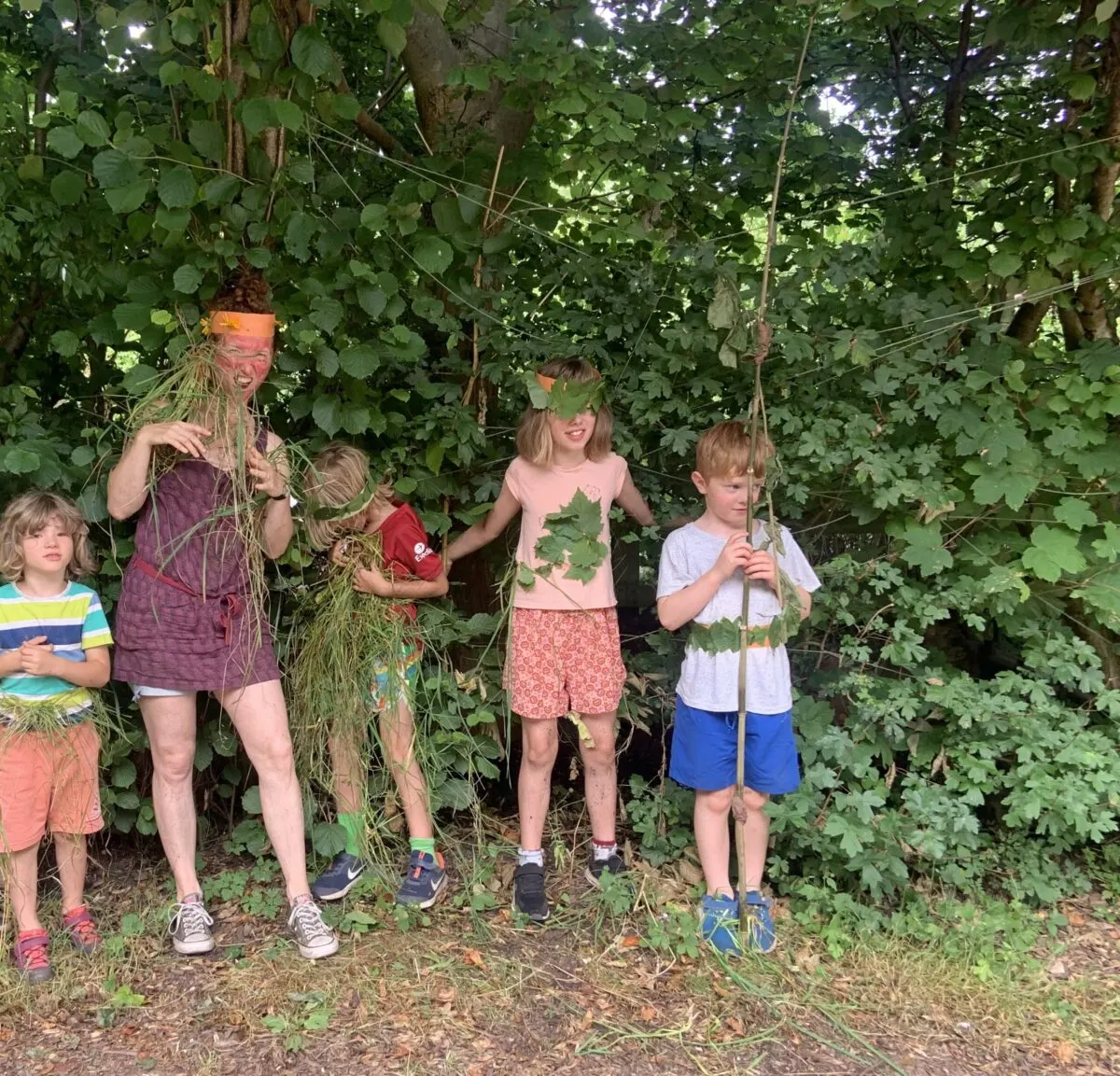 An adult and four children in a dense woodland area holding vines and branches, surrounded by lush green foliage.