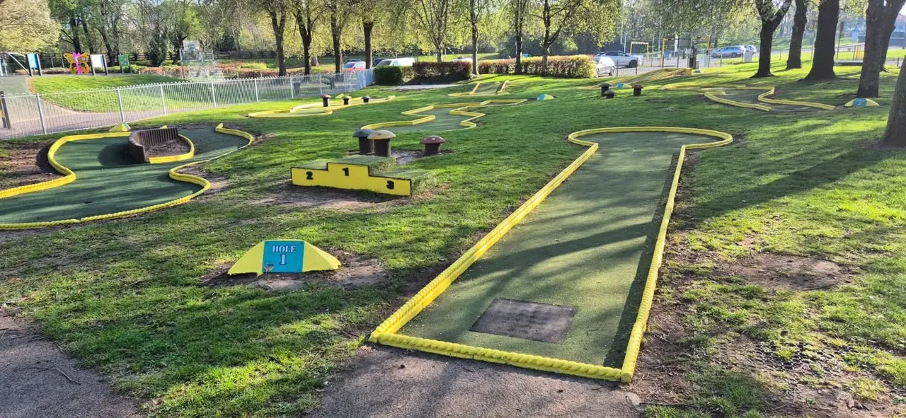 Minigolf course in a park with yellow-bordered green playing surfaces, obstacles and hole markers among trees and grass.