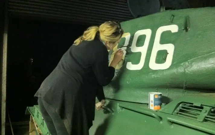 Woman in business suit inspecting the side of a green military tank numbered 96 at night.
