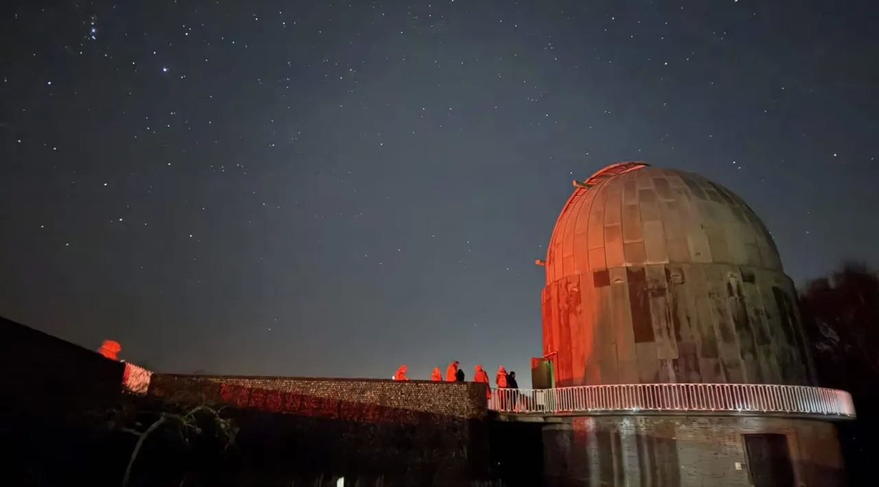 Observatory dome illuminated in red at night with visitors on viewing platform, starry sky above.