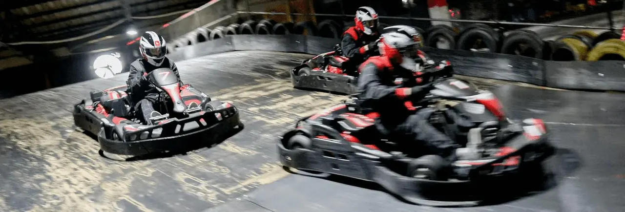 Multiple go-kart racers in helmets and red racing suits competing on an indoor track with tyres lining the barriers.