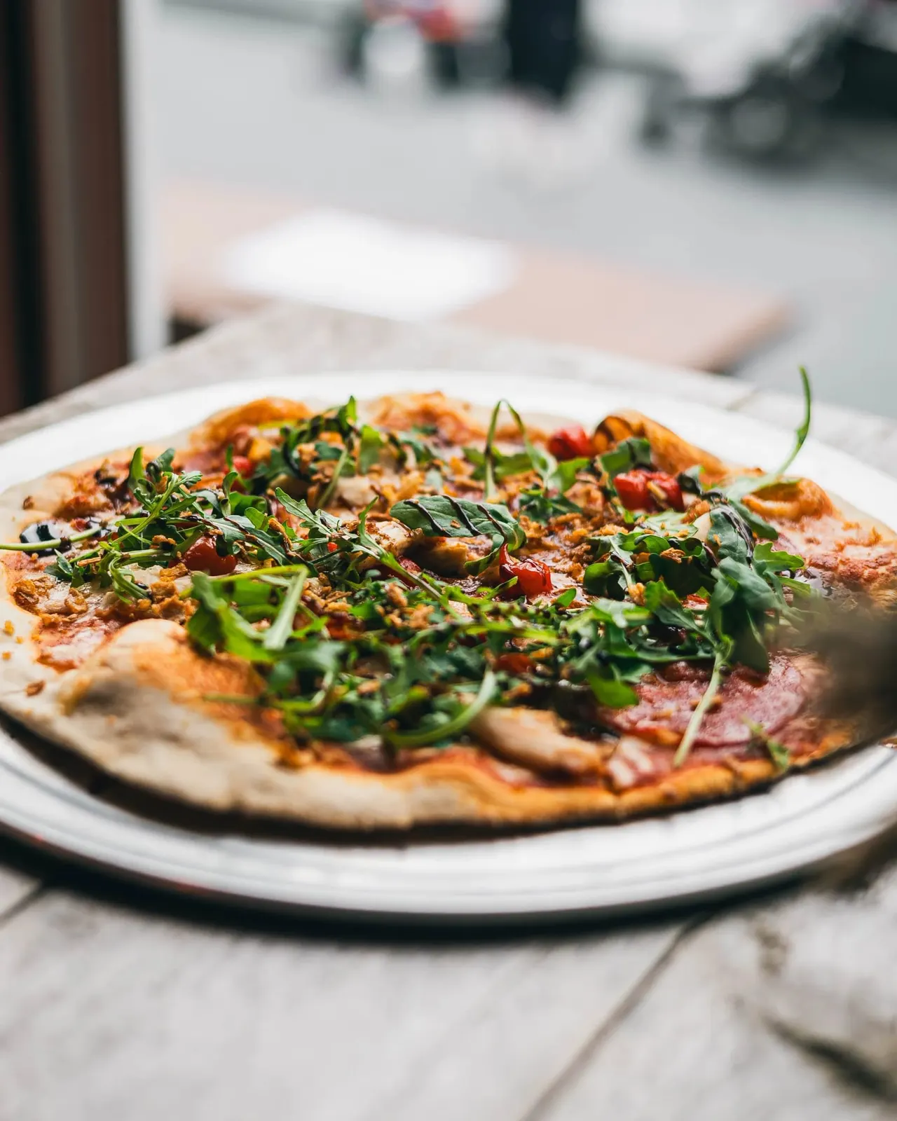 Pizza topped with cured meat, vegetables and fresh rocket leaves served on a white plate outdoors.