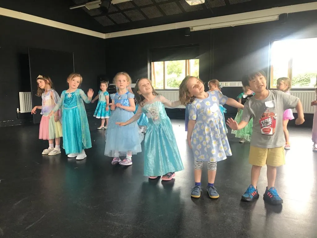 Children aged roughly 5-10 participate in a drama or dance class in a dark studio space, some wearing costume dresses.