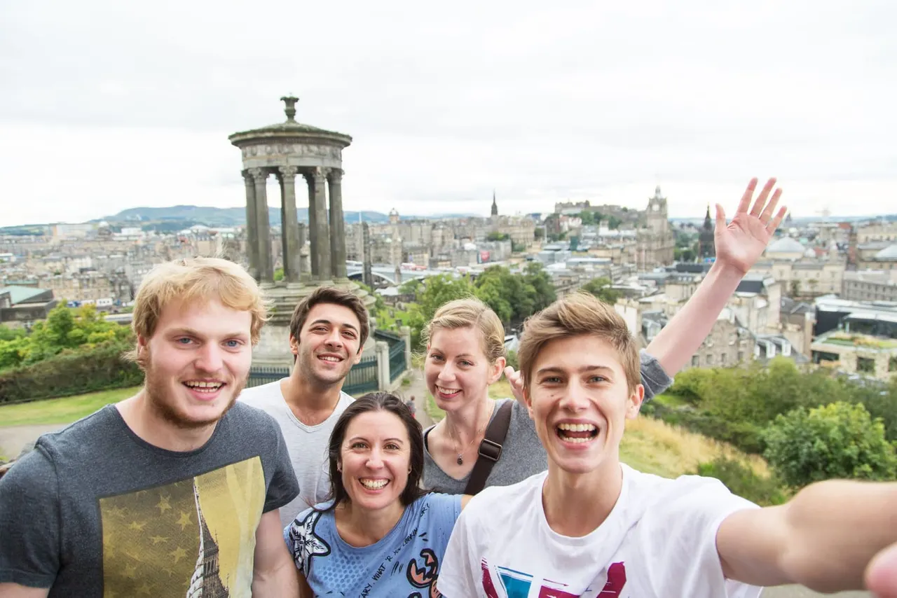 A group of five young people taking a selfie on Calton Hill, with Edinburgh's cityscape and historic monument visible behind them.