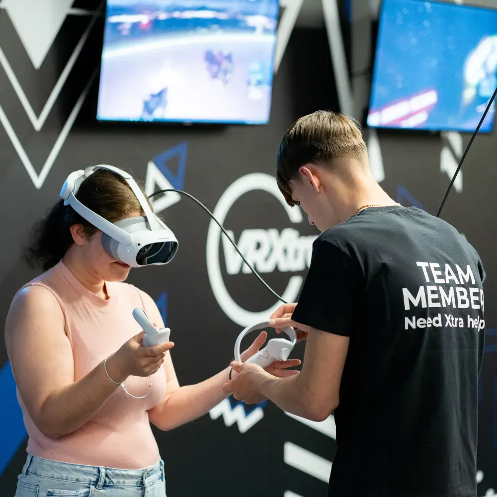 Two people using VR headsets and controllers at an indoor entertainment venue with screens displaying games.