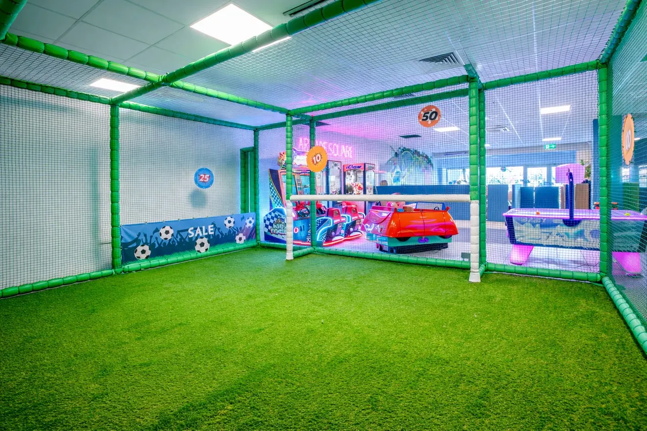 Indoor soft play area with green netting, artificial grass floor, and colourful play equipment including ride-on toys and ball pit games.