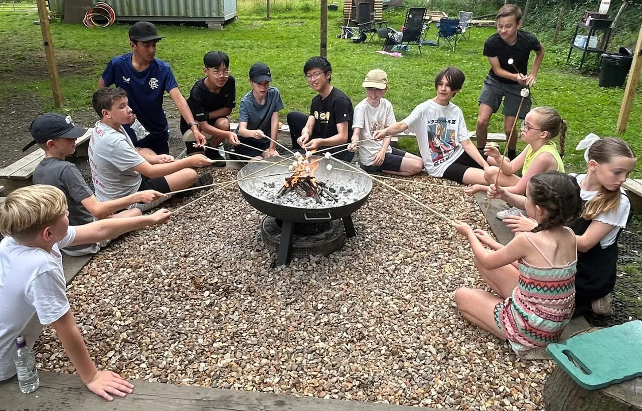 Large group of children and young adults roasting marshmallows around a fire pit outdoors.