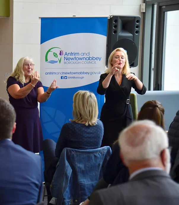 Two women presenting to an audience in front of a blue Antrim and Newtownabbey banner in an indoor venue.