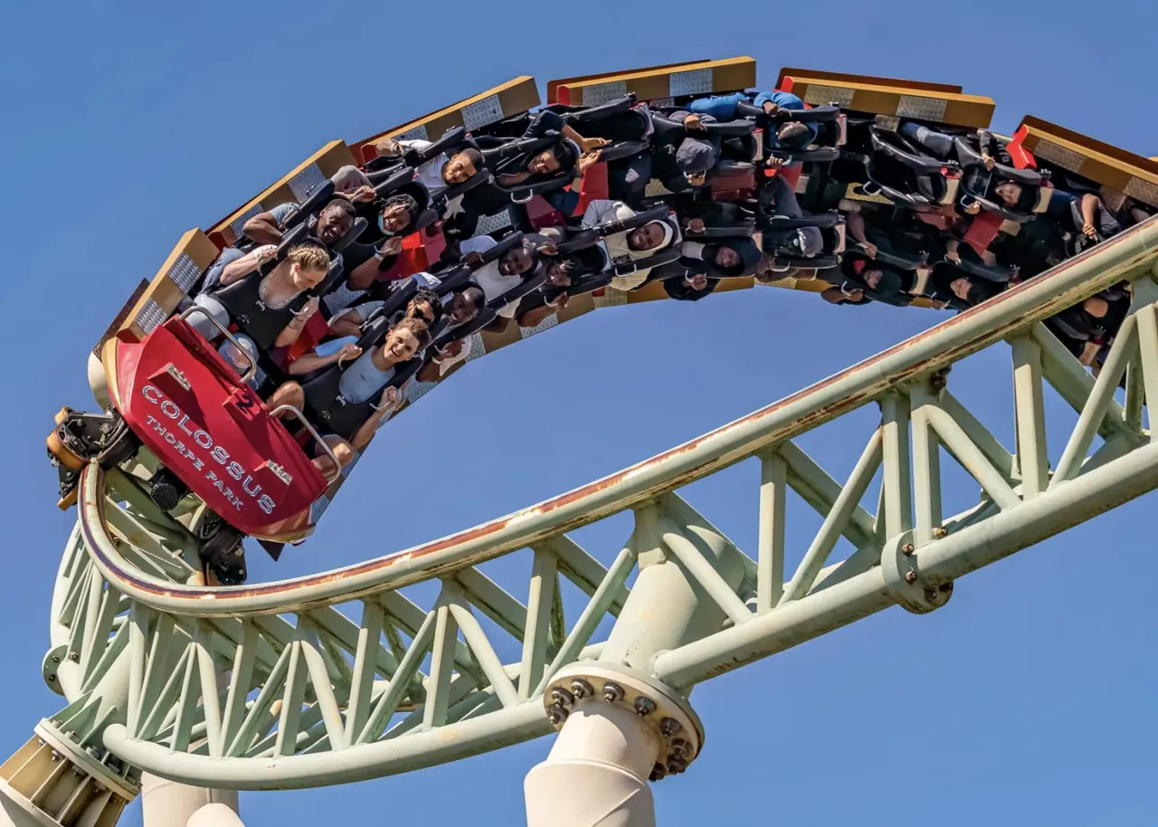 Riders on a red roller coaster car cresting a steep curve with arms raised against a clear blue sky.