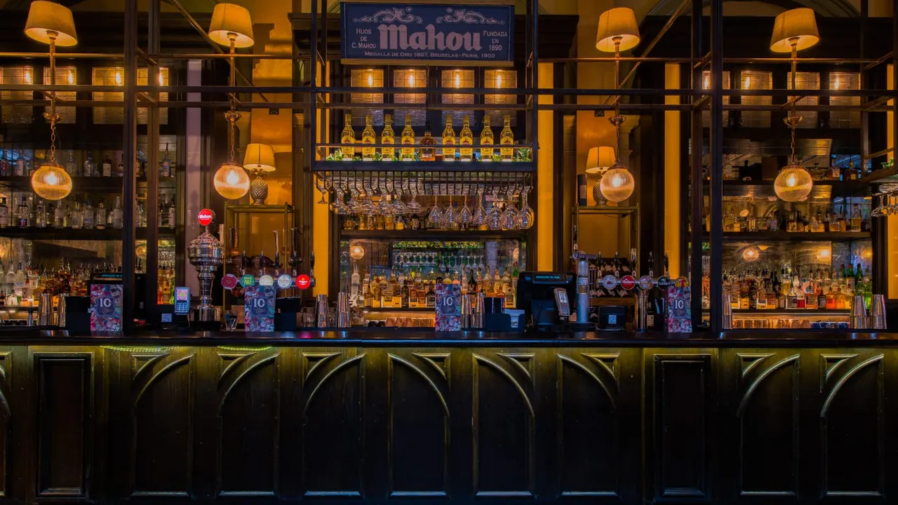 An elegant bar interior with illuminated shelving displaying spirits bottles, vintage signage, hanging amber globe lights, and a dark wooden counter.