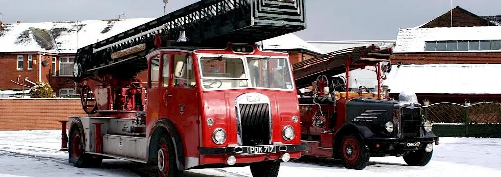 Two vintage red fire engines parked on snowy ground with brick buildings behind.