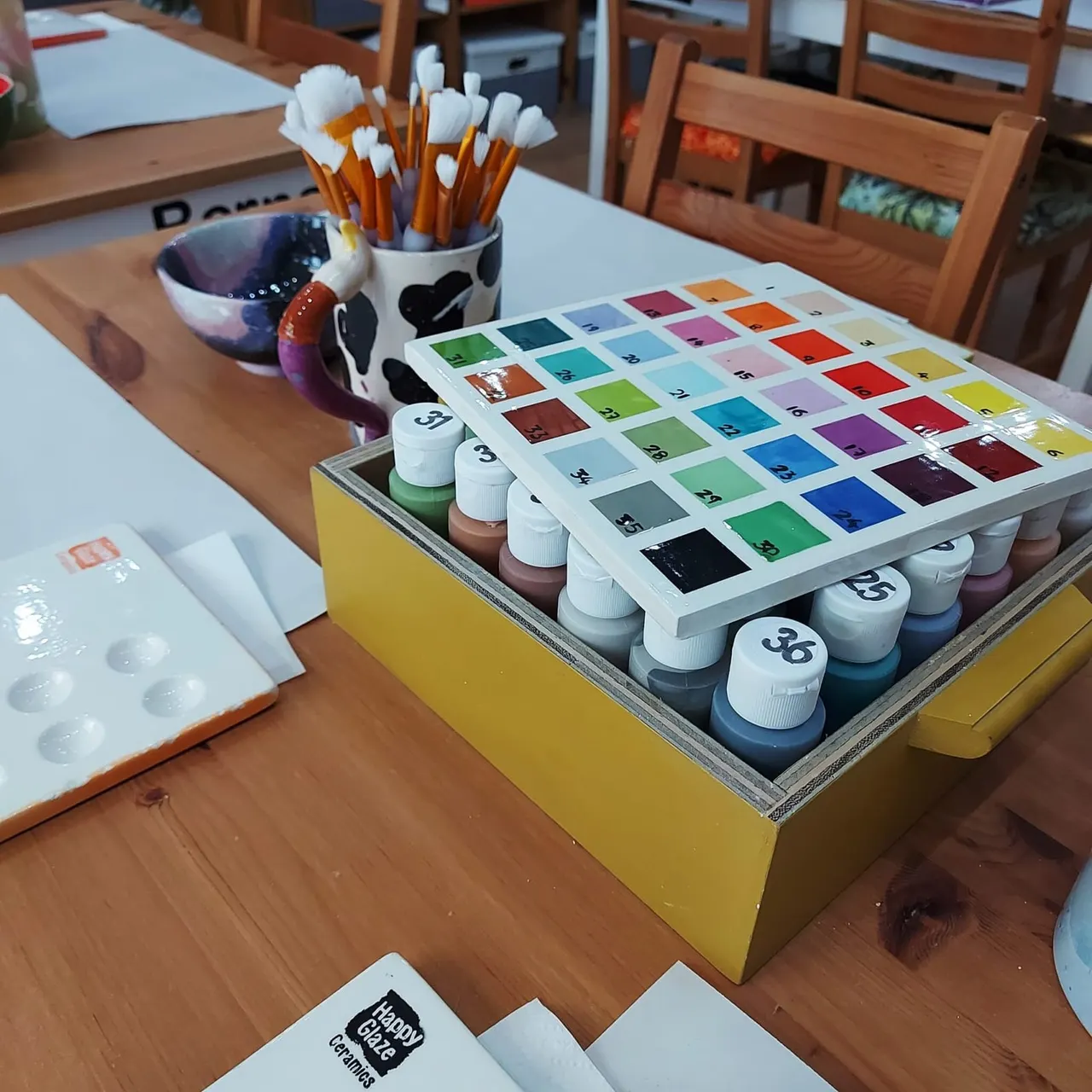 Art activity table with paint pots, brushes, colour swatches and ceramic pieces in a craft studio setting.