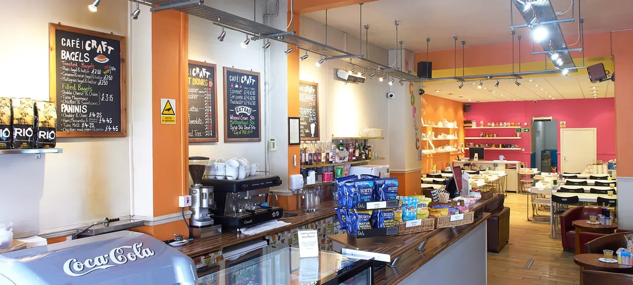 Interior of a café with a service counter, espresso machine, and menu boards, vibrant orange and pink walls, and shelving with craft supplies in the background.