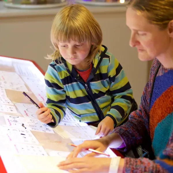 A young child in a striped jacket draws on paper whilst an adult helps guide them at an interactive activity table.