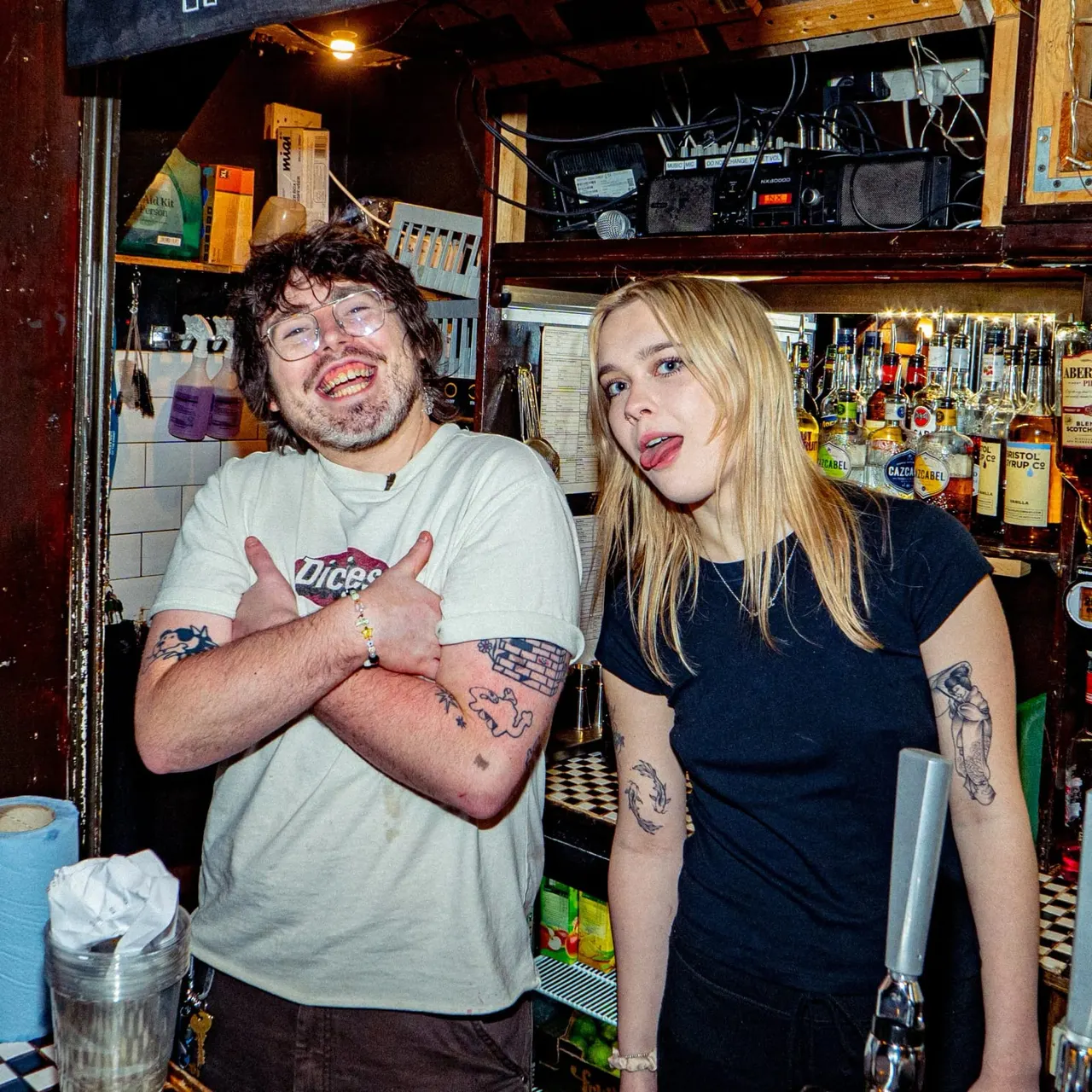 Two people posing playfully behind a bar counter lined with spirit bottles and bar equipment.