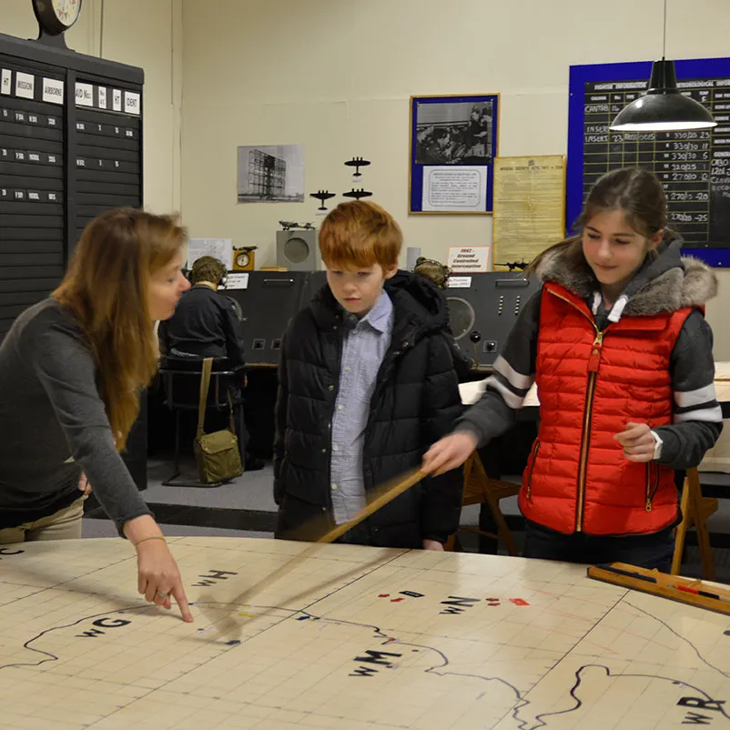 Three young visitors examine a large floor map with a guide in a museum displaying radar equipment and aircraft models.