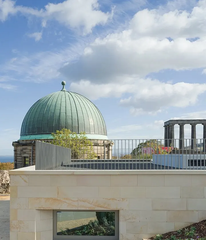 Historic domed building with copper roof and observation platform overlooking coastal landscape under blue sky with white clouds.