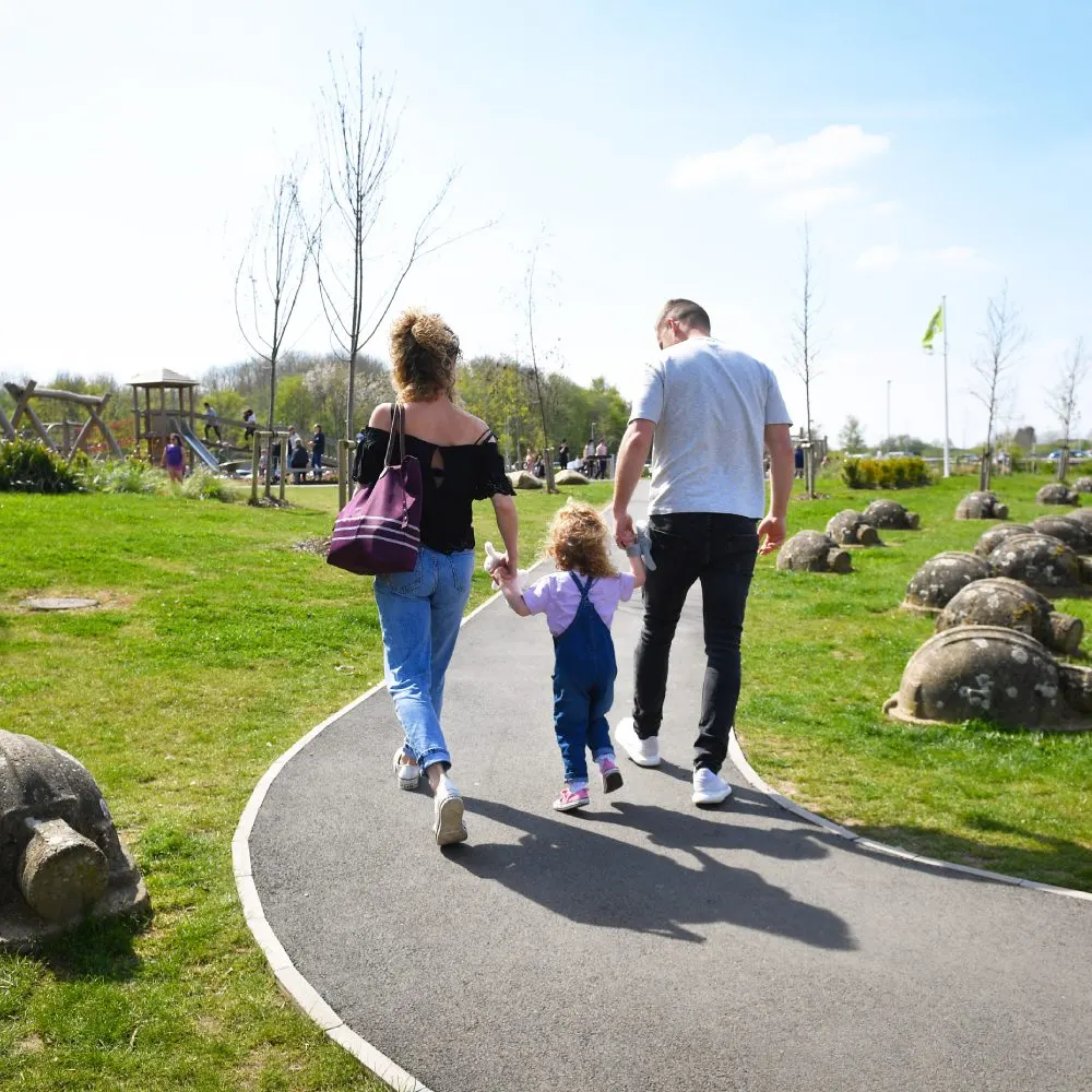 Family walking on a paved path through a park with playground and green spaces visible in the background.