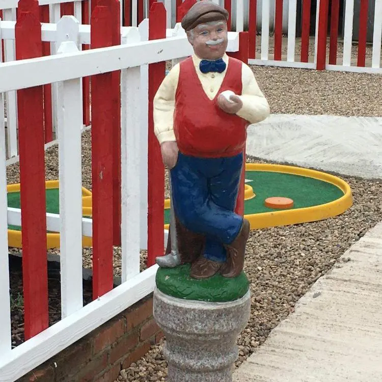 Painted figurine of a man in red waistcoat and blue trousers stands beside a red and white fence on a miniature golf course.