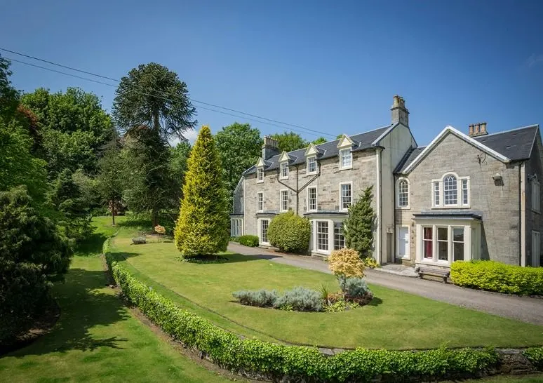 Stone country house with manicured lawns, mature trees, and hedged gardens under blue sky.