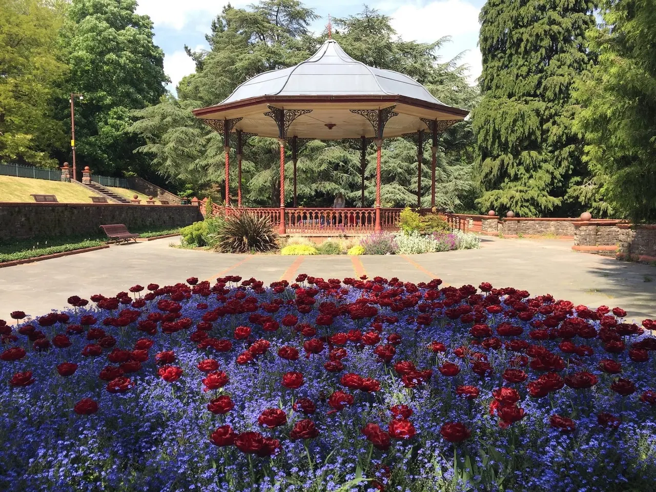 Red and blue flowering plants in foreground with ornate Victorian bandstand, surrounding trees and manicured gardens beyond.
