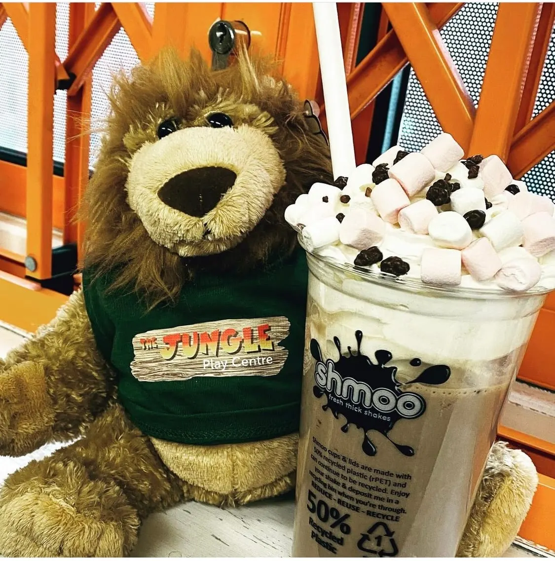 A soft toy lion mascot holding a tall glass of milkshake topped with marshmallows and chocolate pieces.