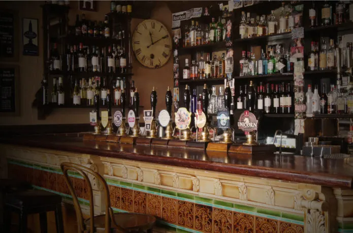 Traditional pub bar counter with multiple beer pumps, shelves of bottles behind, a large clock, and bar stools.