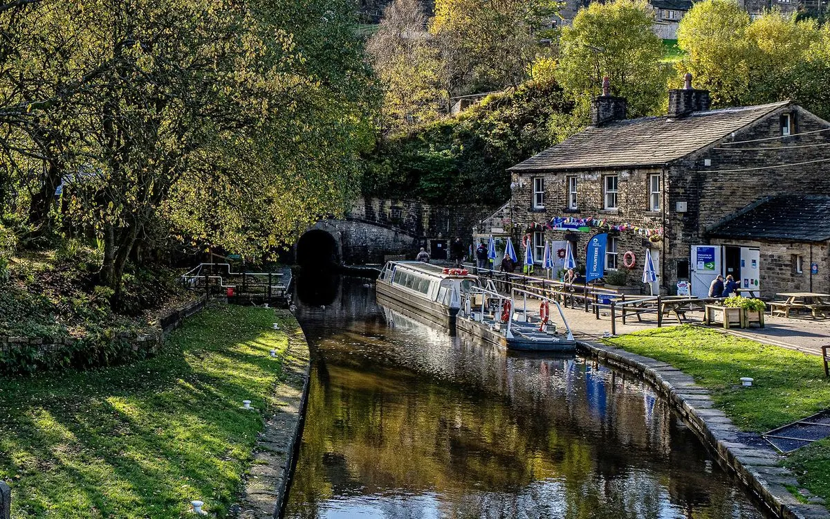 Narrowboat moored alongside a stone building at a canal visitor centre, surrounded by leafy trees and green grass.