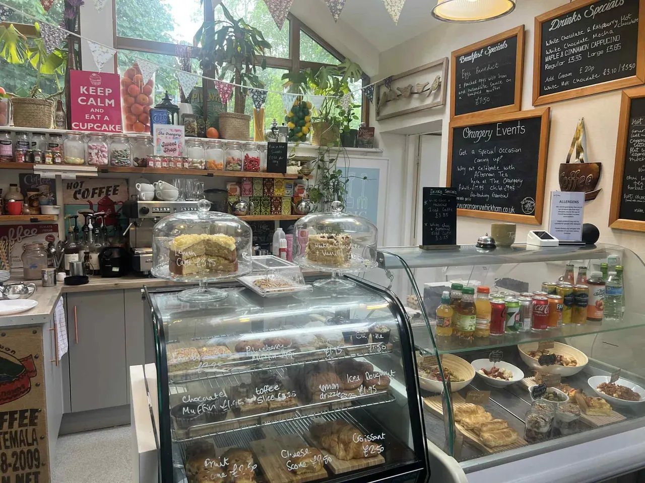 Interior of a café with display counters showing cakes and pastries, coffee machine, hanging plants, and menu boards on the walls.