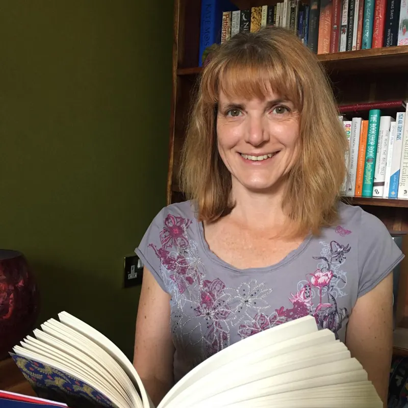 Woman smiling at camera whilst holding an open book, with bookshelves visible behind her.
