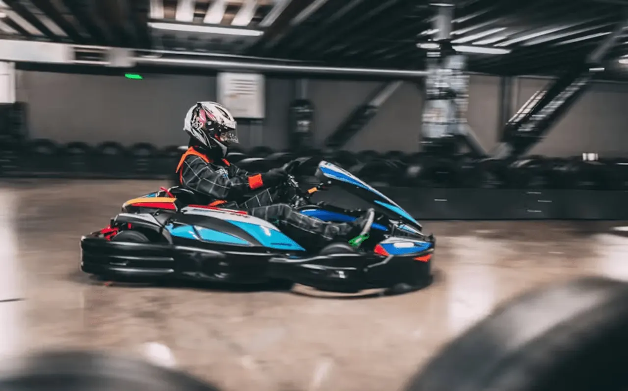 Person in helmet and racing suit driving blue and black go-kart around indoor track.