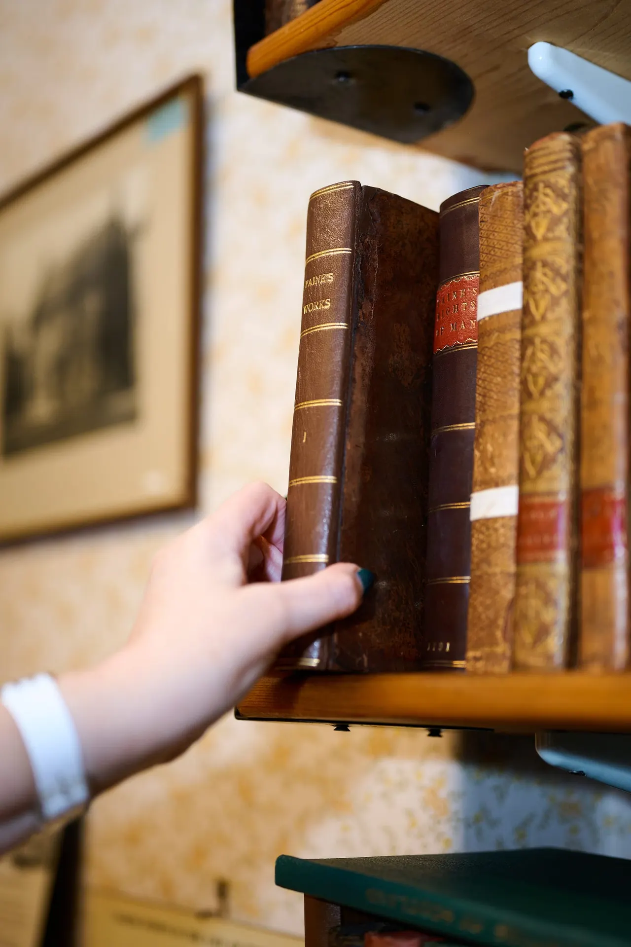Hand pulling an antique leather-bound book from a wooden shelf lined with historic books.