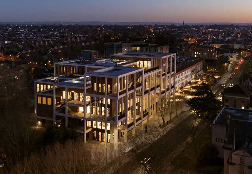 Modern multi-storey building with illuminated windows at dusk, overlooking a residential town sprawling across the landscape.