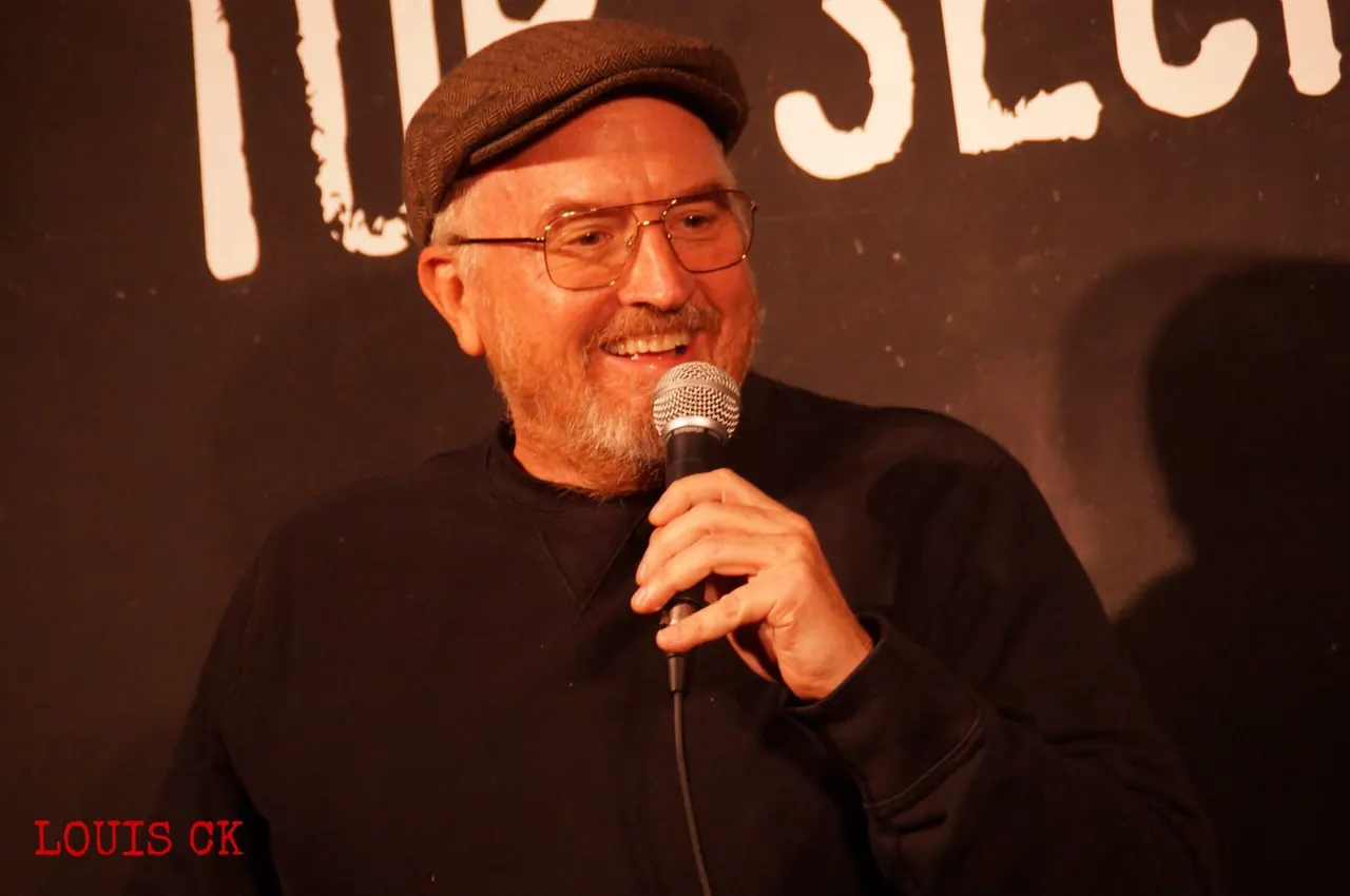 Man wearing brown cap and glasses speaking into microphone on stage at comedy club.