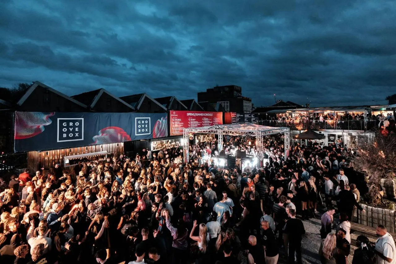 Large crowd of people dancing and celebrating at an outdoor music event with illuminated stages and branding at dusk.