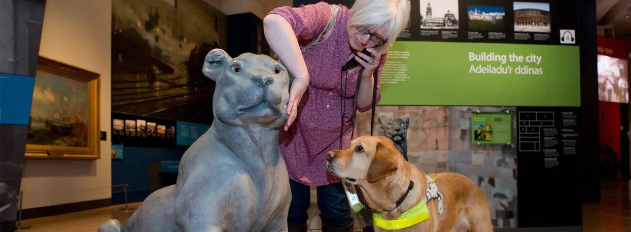 Woman petting a hippo sculpture while a service dog looks on inside a museum exhibition space.