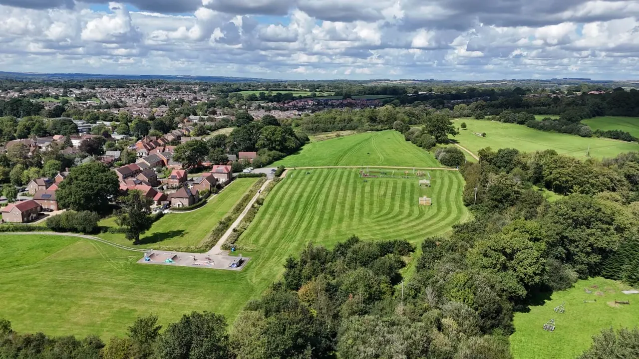 Aerial view of rolling green fields with a small play area beside a residential suburb under cloudy sky.